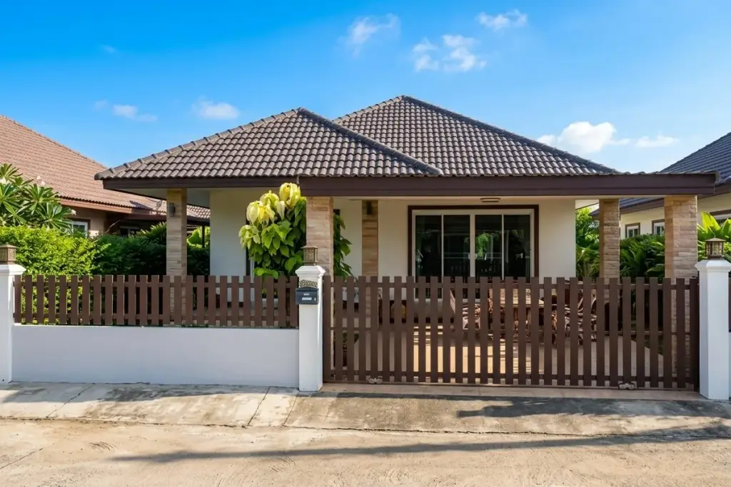 Petchlada 3: Exterior view of a house with a brown picket fence, tiled roof, and lush green hedges.
