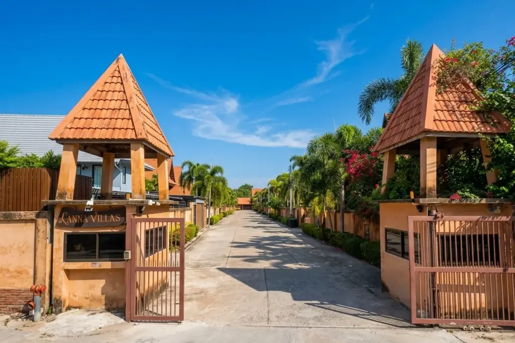 Lanna Villa entrance gate with two gatehouses, a paved road, and lush palm trees leading into the property.