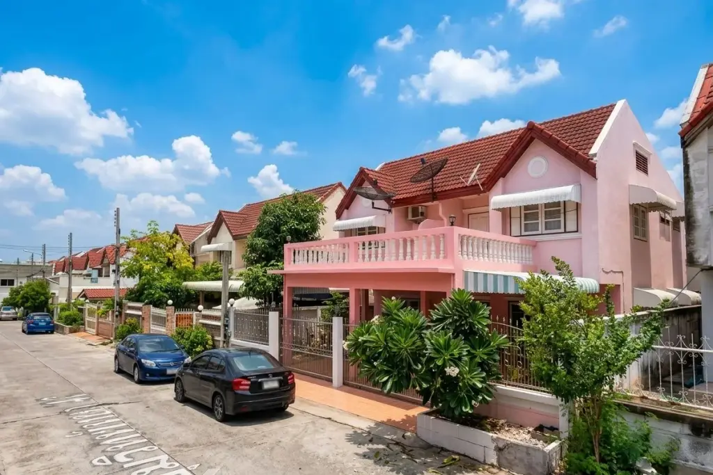 Sabaijai Village street view with a pink house, red tiled roof, balcony, and parked cars under a blue sky.