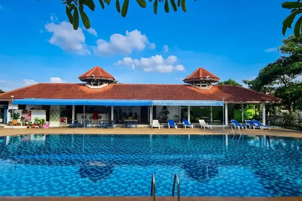 Chokchai Village 4 large outdoor swimming pool with blue tiles and a clubhouse building with a red roof.