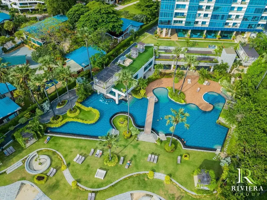 Palm Wongamat aerial view of a large, blue swimming pool with a bridge, surrounded by green lawns, palm trees, and sun loungers.