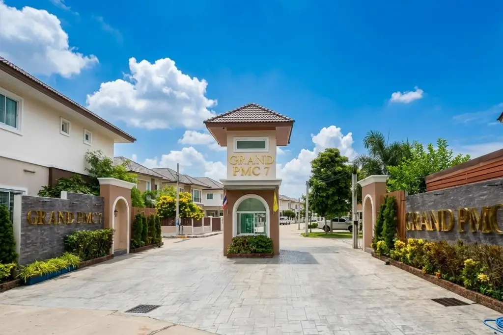 Grand PMC 7 main entrance with security booth, barrier gate, and residential buildings under a blue sky.