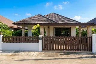 Petchlada 3: Exterior view of a house with a brown picket fence, tiled roof, and lush green hedges.