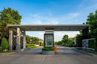 Life @ Banglamung security gate entrance with guardhouse, automatic barriers, and landscaping under a cloudy sky