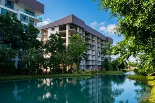 The Park Jomtien. Modern condo building under construction next to a serene pond with lush green trees and blue sky.