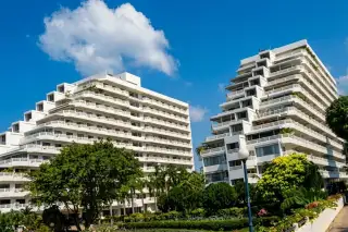 Jomtien Condotel & Village exterior view of the white terraced building with multiple balconies and surrounding lush green trees.