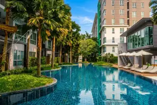 Natureza Art outdoor swimming pool with blue tiles, wooden deck, lounge chairs, and a person relaxing, surrounded by lush greenery and modern condo buildings.