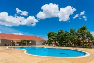 Chokchai Garden Home 2 large outdoor swimming pool with surrounding patio, lounge chairs, and buildings under a blue sky.