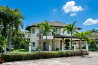 Green Field Villas 1 two-story house with light facade, dark roof, palm trees, green lawn, and fence under a blue sky.