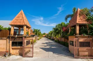 Lanna Villa entrance gate with two gatehouses, a paved road, and lush palm trees leading into the property.