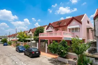 Sabaijai Village street view with a pink house, red tiled roof, balcony, and parked cars under a blue sky.