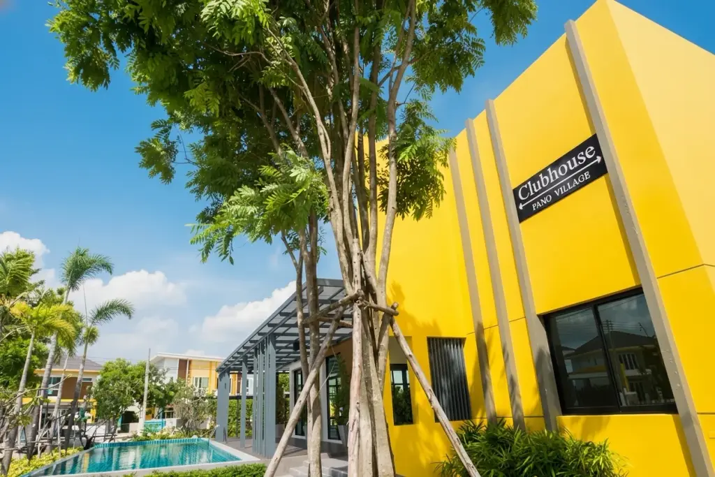Pano Village bright yellow clubhouse building with sign, adjacent swimming pool, and lush green trees under a blue sky.