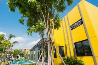 Pano Village bright yellow clubhouse building with sign, adjacent swimming pool, and lush green trees under a blue sky.