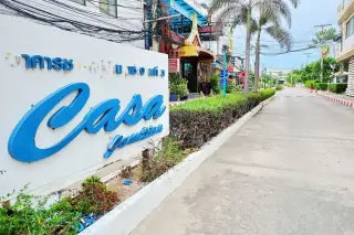 Casa Jomtien entrance sign with blue lettering, lush green hedges, and a paved road leading into the community.