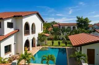 Mabprachan Gardens outdoor swimming pool with surrounding white villa, green lawn, and tropical plants under a blue sky.
