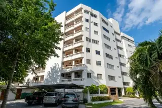 Ruamchok Condo View 2: Exterior view of the white multi-story condo building with balconies, shaded parking, and lush trees.