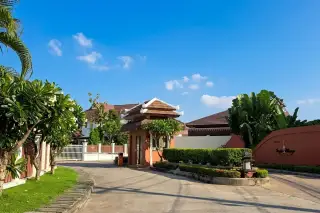 Tropical Village entrance with a traditional Thai-style gatehouse, paved road, green lawns, and tropical plants.