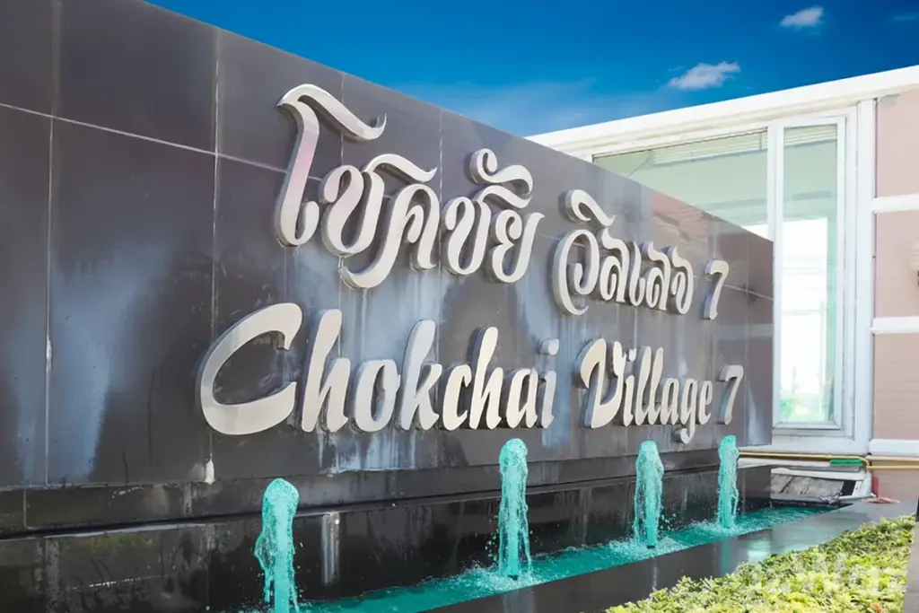 Chokchai Village 7 entrance sign in silver lettering on black, with small water fountains in front.