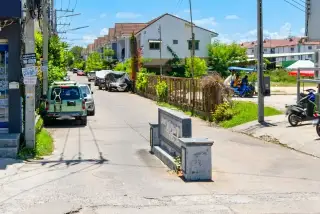 Moo Baan Chawala entrance sign on a paved street with parked cars, houses, and power lines under a sunny sky.