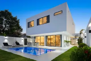 D-Sign Homes modern white house with a rectangular swimming pool, green lawn, lounge chairs, and outdoor dining set.
