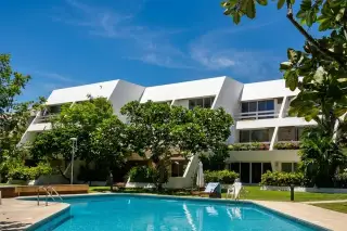 Bang Saray Beach Condominium exterior with a clear blue swimming pool, lush green trees, and white building facade under a bright sky.
