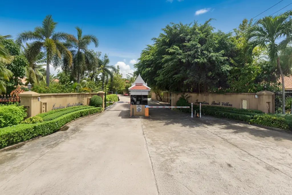 Jomtien Park Villas entrance gate with security booth, lush greenery, and palm trees under a cloudy sky.