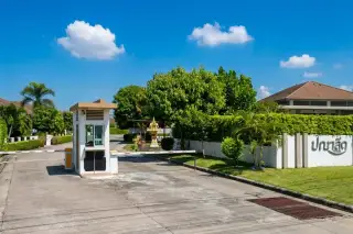 Panalee Village main entrance with a security gatehouse, barrier arm, paved road, and green landscaping under a blue sky.
