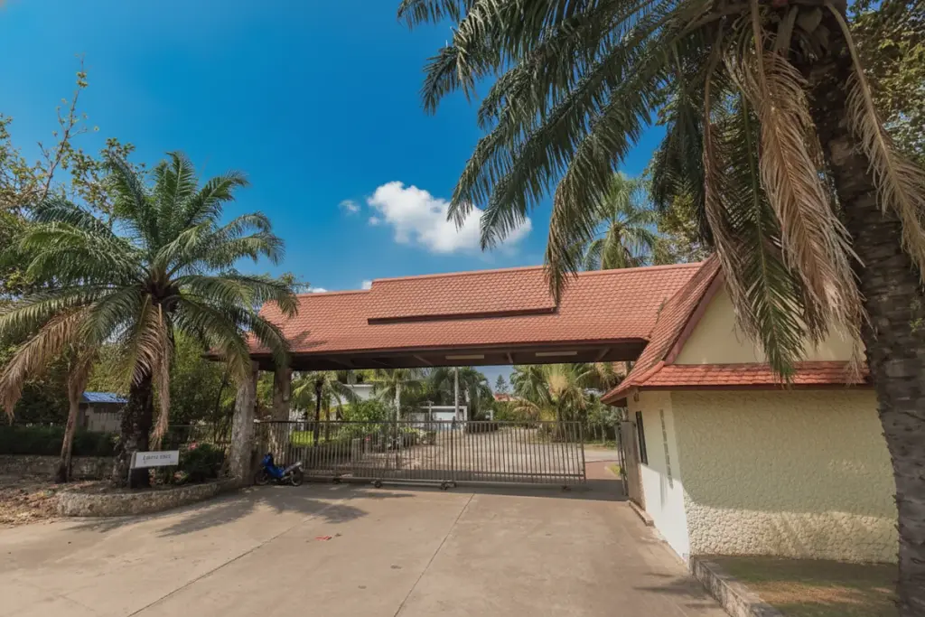 Little Hill Village entrance gate with a large archway, red tiled roof, gatehouse, and lush palm trees.