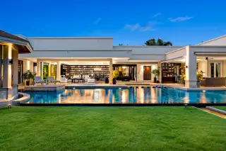 The Vineyard luxurious house exterior at dusk with a large swimming pool, illuminated living areas, and green lawn.
