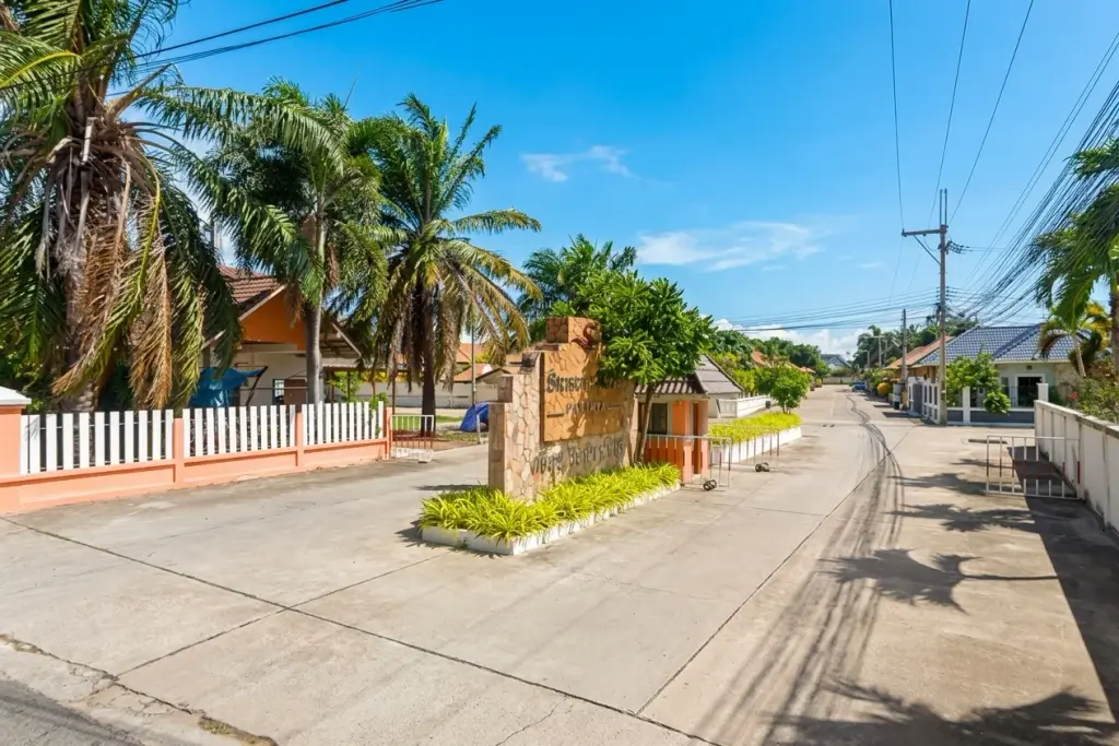 Srisuk Villa entrance gate with stone sign, palm trees, and residential road under a blue sky in Pattaya, Thailand.