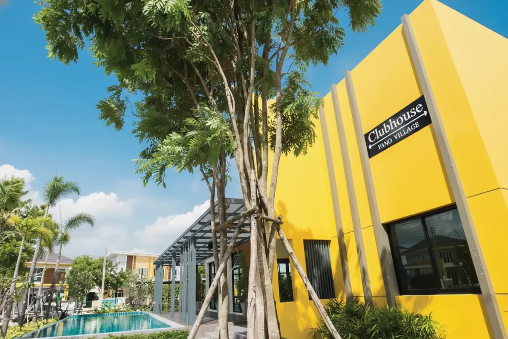 Pano Village bright yellow clubhouse building with sign, adjacent swimming pool, and lush green trees under a blue sky.