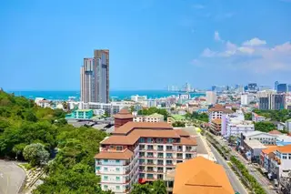 Estanan Condo: Panoramic view of Pattaya cityscape with tall buildings, lush green hills, and the ocean under a blue sky.