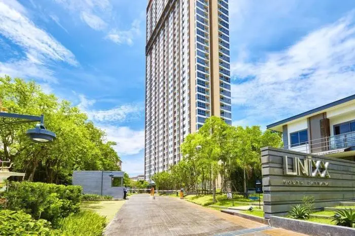 Unixx South Pattaya tall condo building exterior with entrance gate, lush greenery, and property sign on a sunny day.