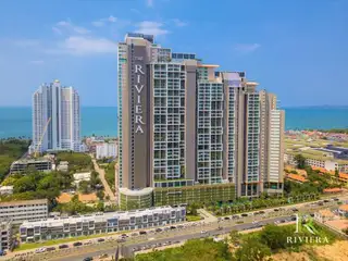 The Riviera Jomtien full exterior view of the tall condo building with 'THE RIVIERA' signage, facing a busy road and the ocean.