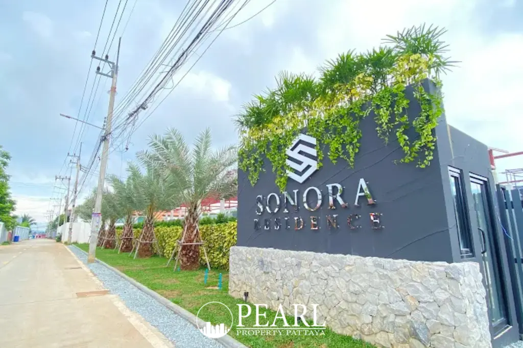 Sonora Residence entrance sign with dark gray background, silver letters, green foliage, and a stone base, beside a paved road with palm trees.