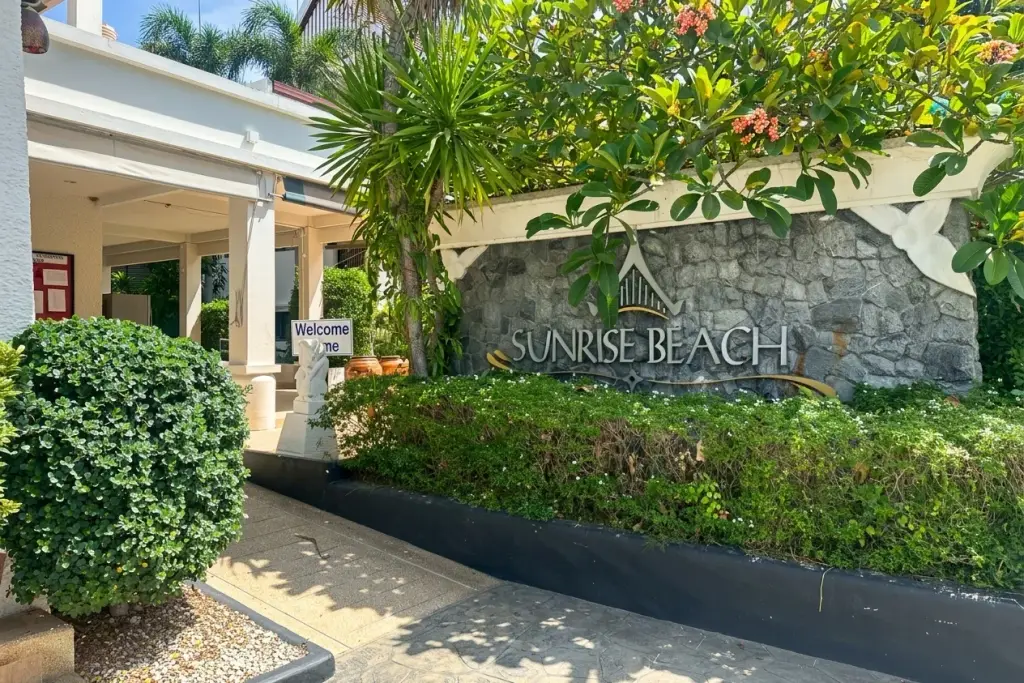 Sunrise Beach Residence stone entrance sign surrounded by lush green foliage and tropical plants on a sunny day.