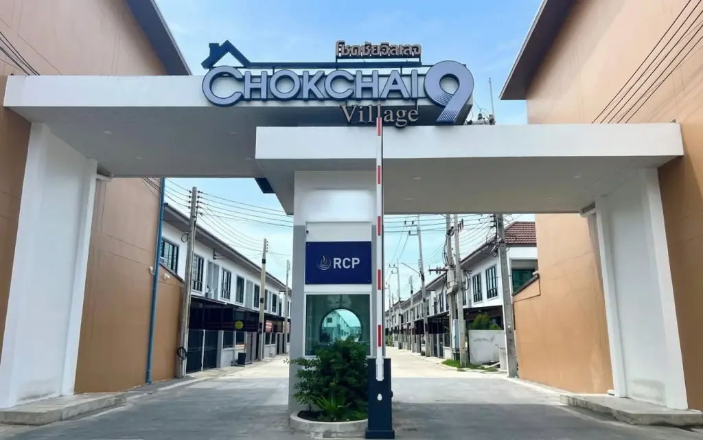 Chokchai 9 village entrance gate with a security barrier, sign, and view of houses along the street.