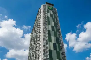 Unicca tall modern condominium building with green and grey facade, adjacent to a multi-lane road with cars and trees under a blue sky.