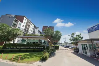 The Trust Central Pattaya condo building exterior with entrance, security gate, and lush landscaping under a bright sky.
