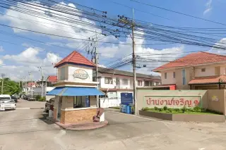 Baan Ngam Charoen 9 entrance with a guardhouse, security barrier, and community sign under a blue sky.