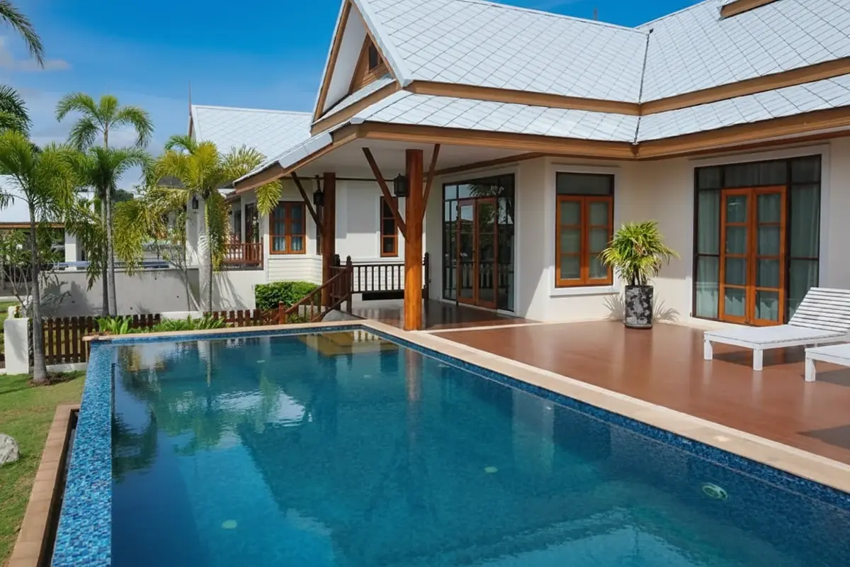 Amorn Village house exterior with a rectangular swimming pool, wooden deck, and white tiled roof under a blue sky.
