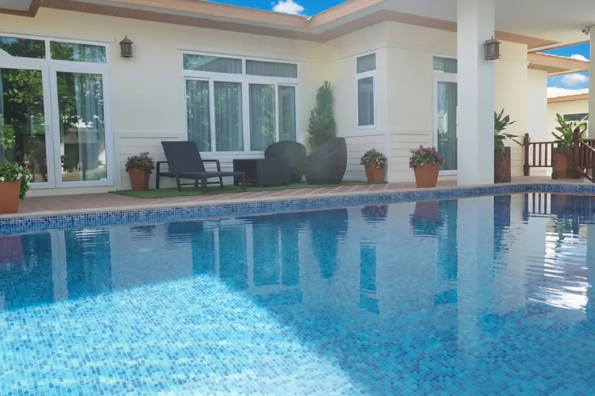 Amorn Village house exterior with a swimming pool reflecting the sky, a patio with seating, and large windows.