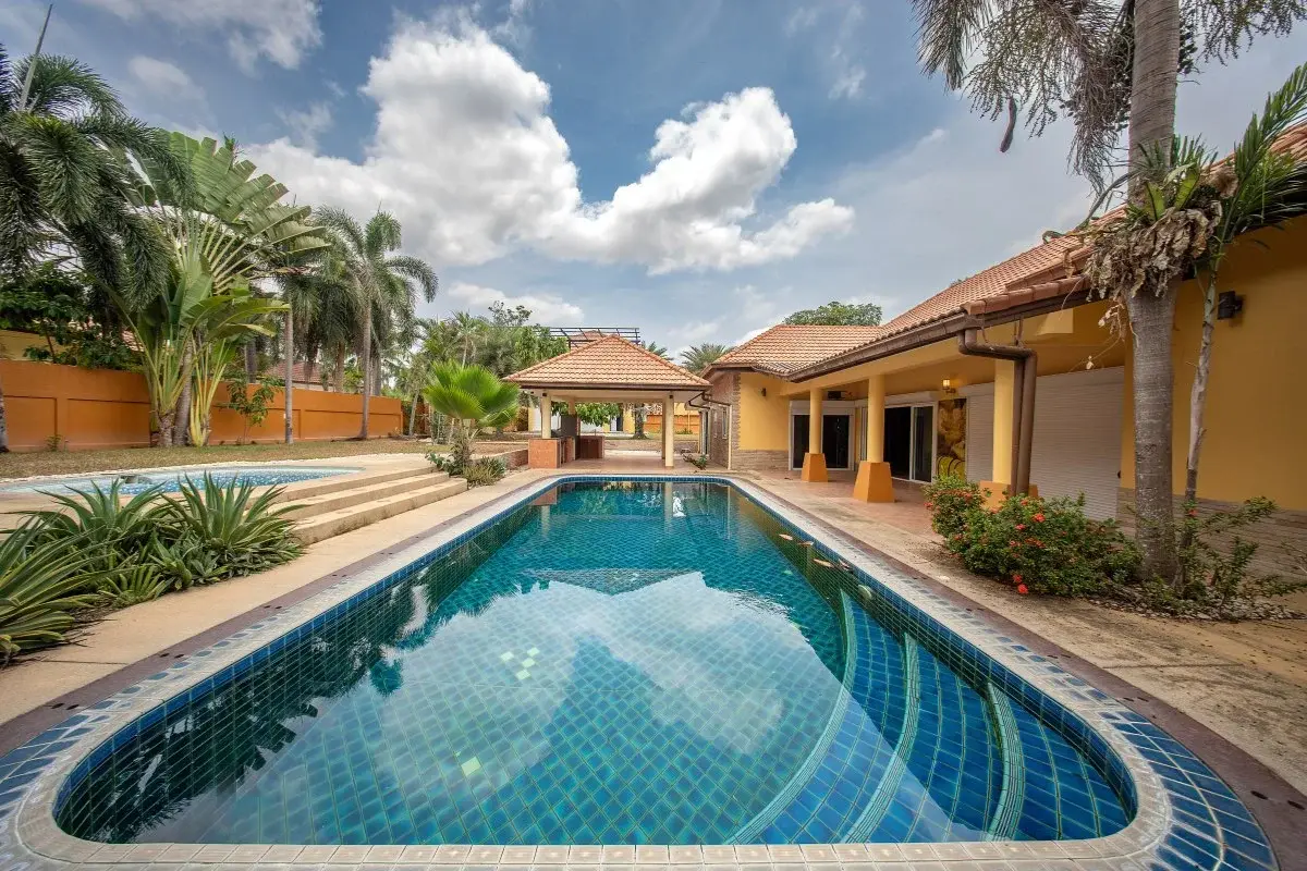 5 Bedroom House for Sale in Huai Yai featuring a long swimming pool, covered patio, and lush palm trees.