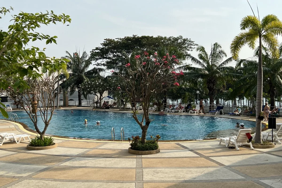 View Talay 7 outdoor swimming pool with sunbathers, palm trees, and tropical plants under a clear sky.