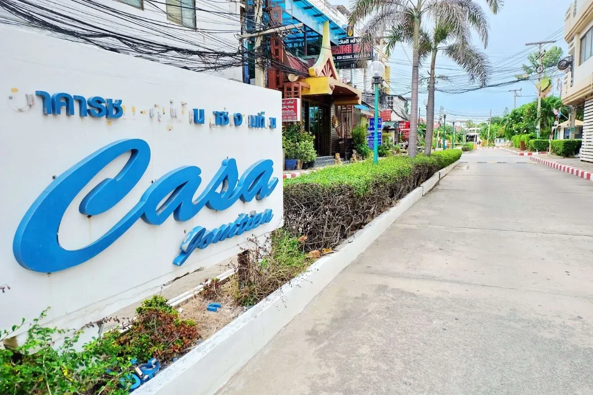 Casa Jomtien entrance sign with blue lettering, lush green hedges, and a paved road leading into the community.