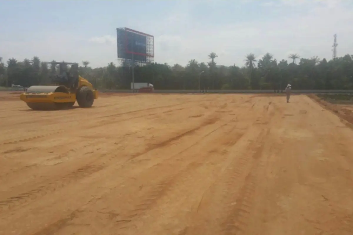 Land for Sale in Nong Pla Lai, wide view of a cleared dirt plot with a road roller and a person, trees, and highway.