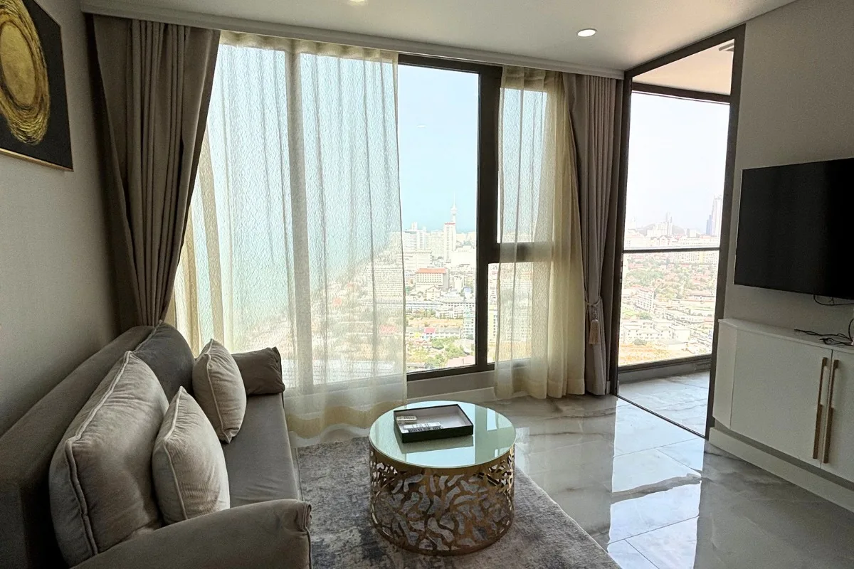 1 Bedroom Condo for Sale & Rent at Copacabana Beach Jomtien: Living room with sofa, coffee table, and large windows with city view.
