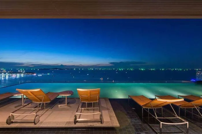 Centric Sea infinity pool with wooden lounge chairs at night overlooking the illuminated ocean.