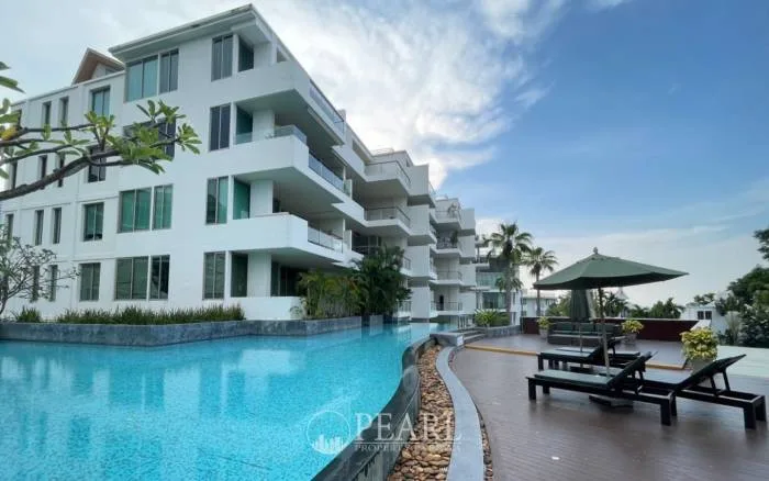 The Sanctuary Wongamat. Modern white condo building with balconies, a curved swimming pool, and sun loungers on a wooden deck.