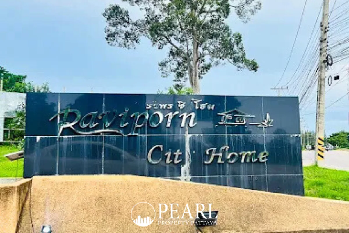 Raviporn City Home gated entrance with guard house, boom barriers, and community sign under a cloudy sky.